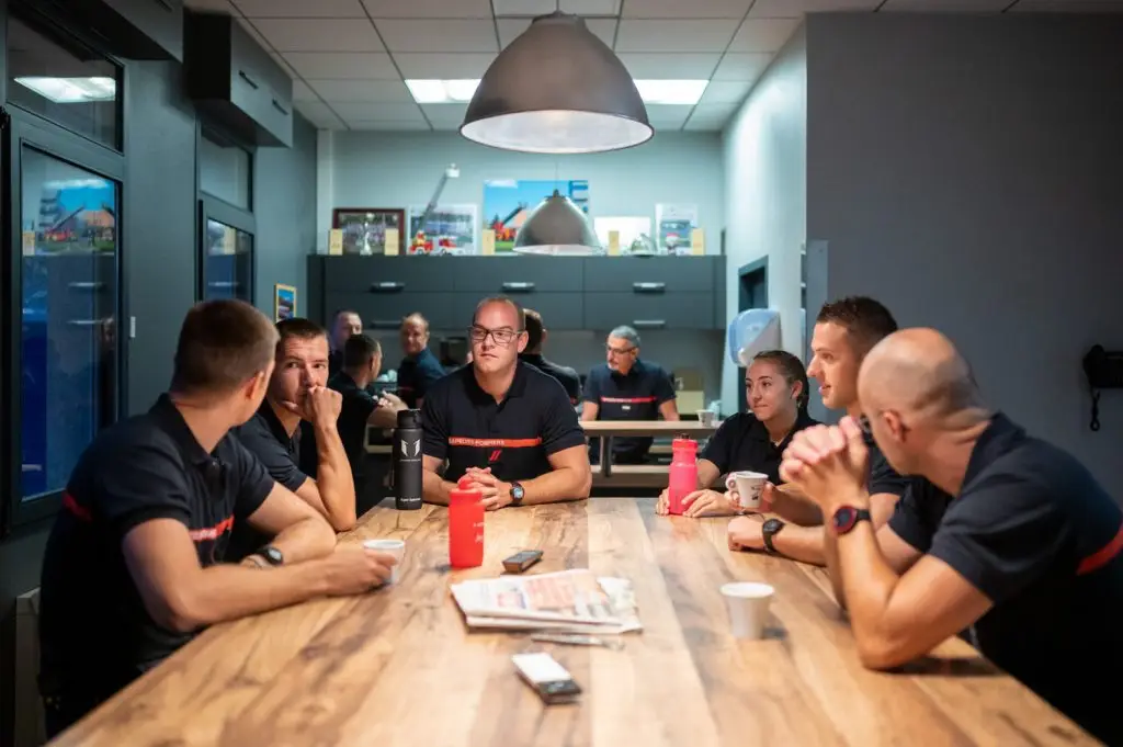 Un groupe de sapeurs-pompiers avec une jeune femme échange autour d'une grande table en bois en caserne, par Vincent Delesvaux, photographe et vidéaste à Lyon.